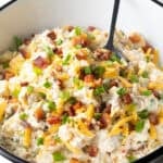A loaded baked potato salad recipe topped with bacon, cheese, and chives, sitting in white bowl with a spoon.
