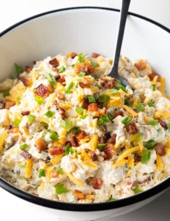A loaded baked potato salad recipe topped with bacon, cheese, and chives, sitting in white bowl with a spoon.