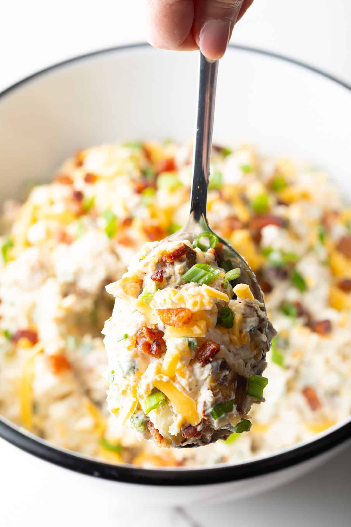 A spoon holding a scoop of loaded baked potato salad over a bowl filled with the salad.