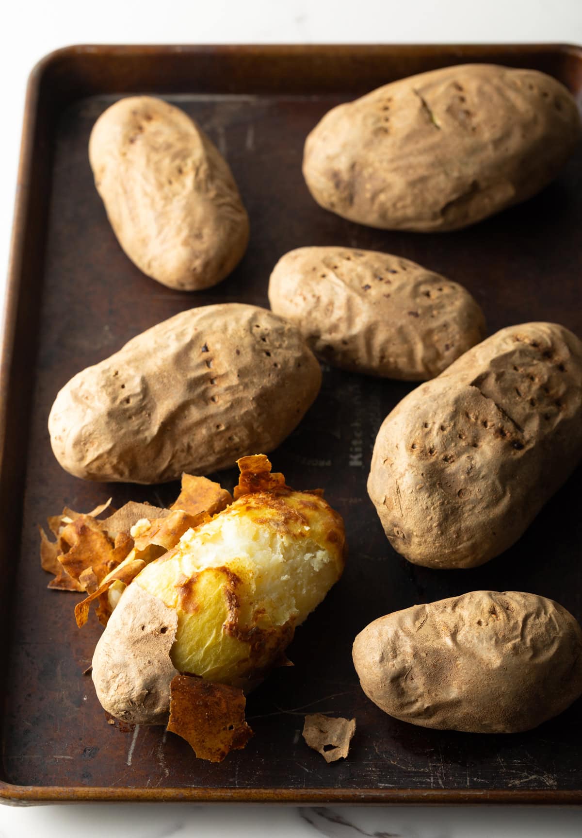 Baked potatoes on a baking sheet, one of which is mostly peeled.