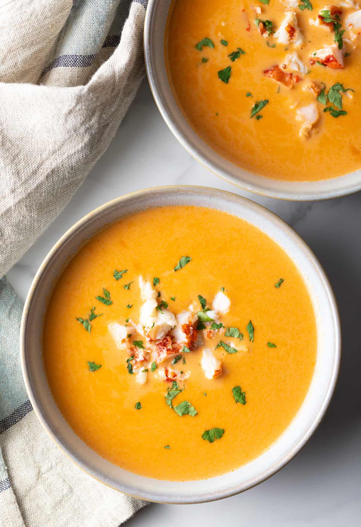 An overhead shot of 2 bowls of a creamy lobster bisque recipe topped with lobster chunks and parsley.