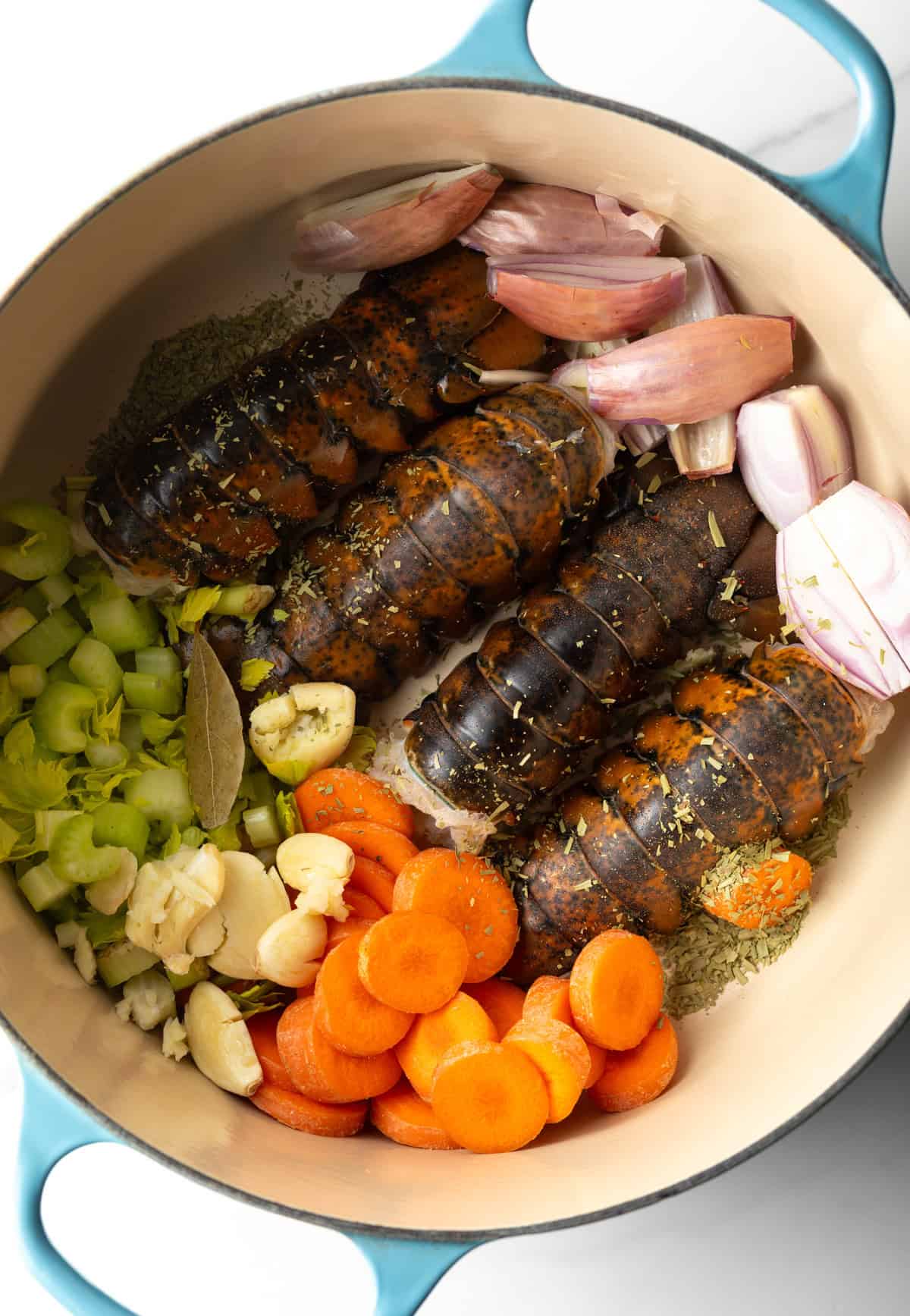 An overhead shot of a pot filled with uncooked lobster tails, carrots, celery, garlic, and shallots.