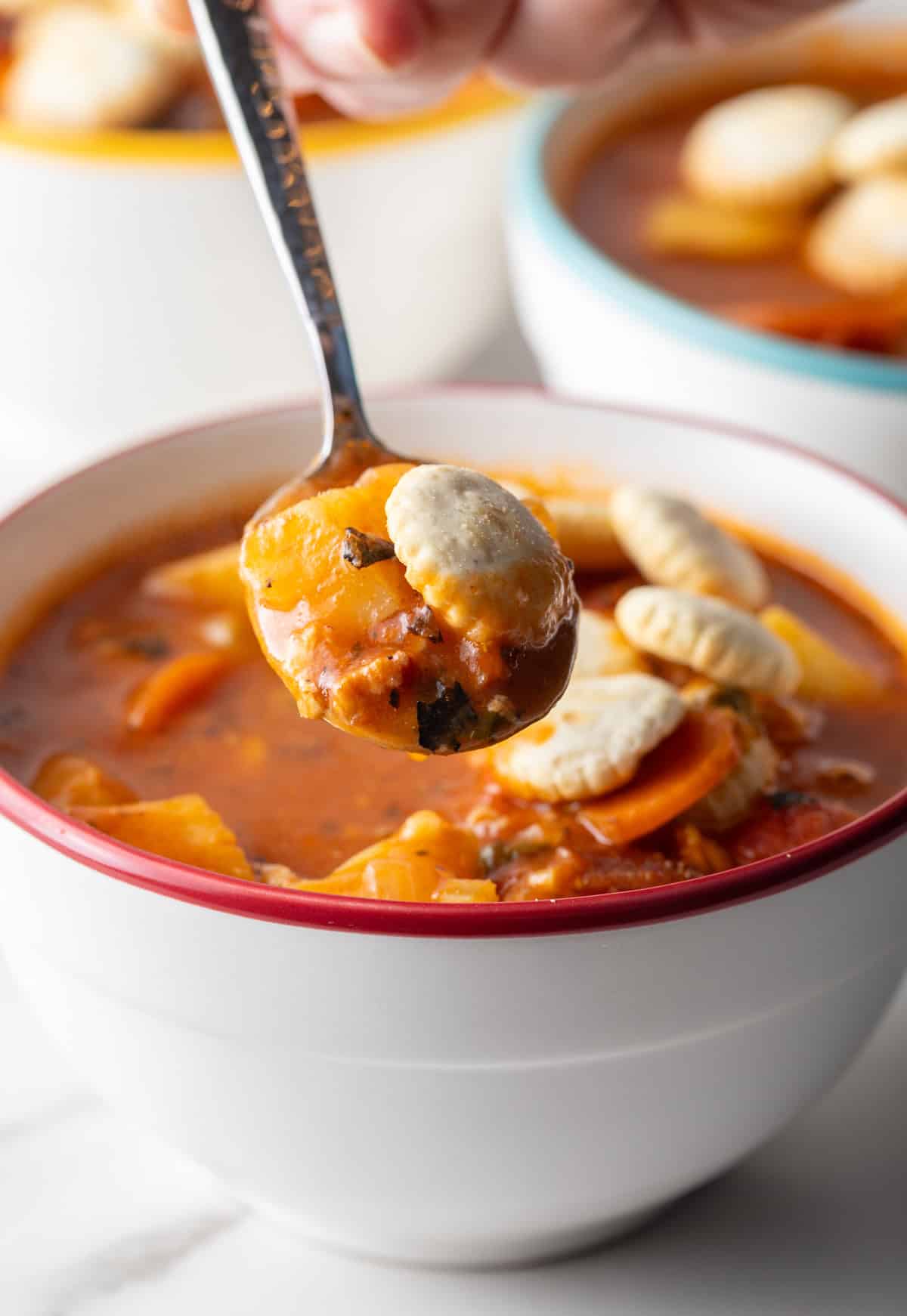 A metal spoon is holding a bite of Manhattan clam chowder with oyster crackers over a white bowl filled with the soup.