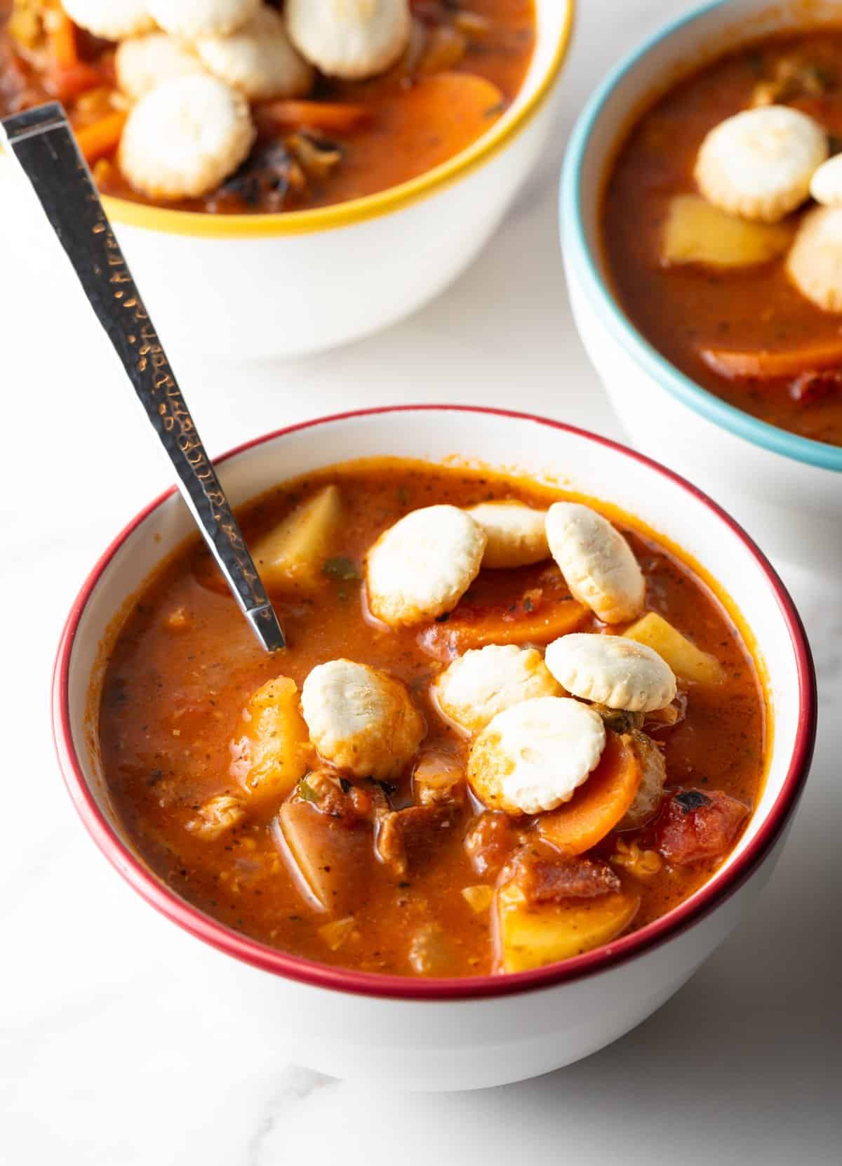 A metal spoon is resting in a bowl of clam chowder Manhattan-style, topped with oyster crackers.