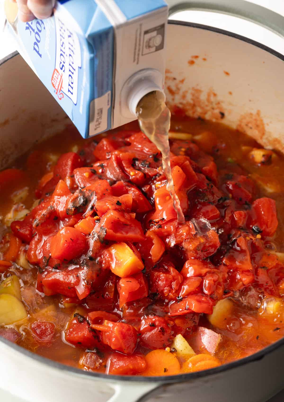 Seafood stock is being poured over a pot filled with vegetables and canned tomatoes.