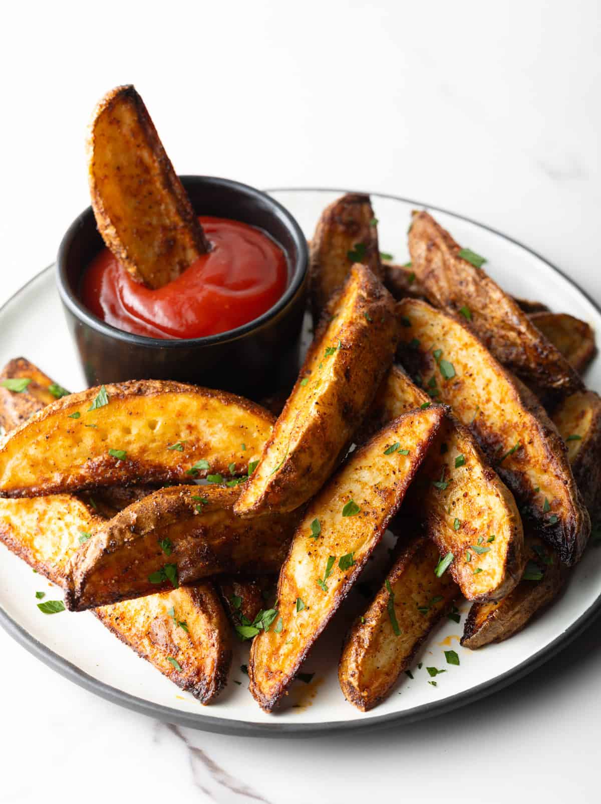 A plate of oven baked potato wedges, one of the wedges is in a ramekin of ketchup.