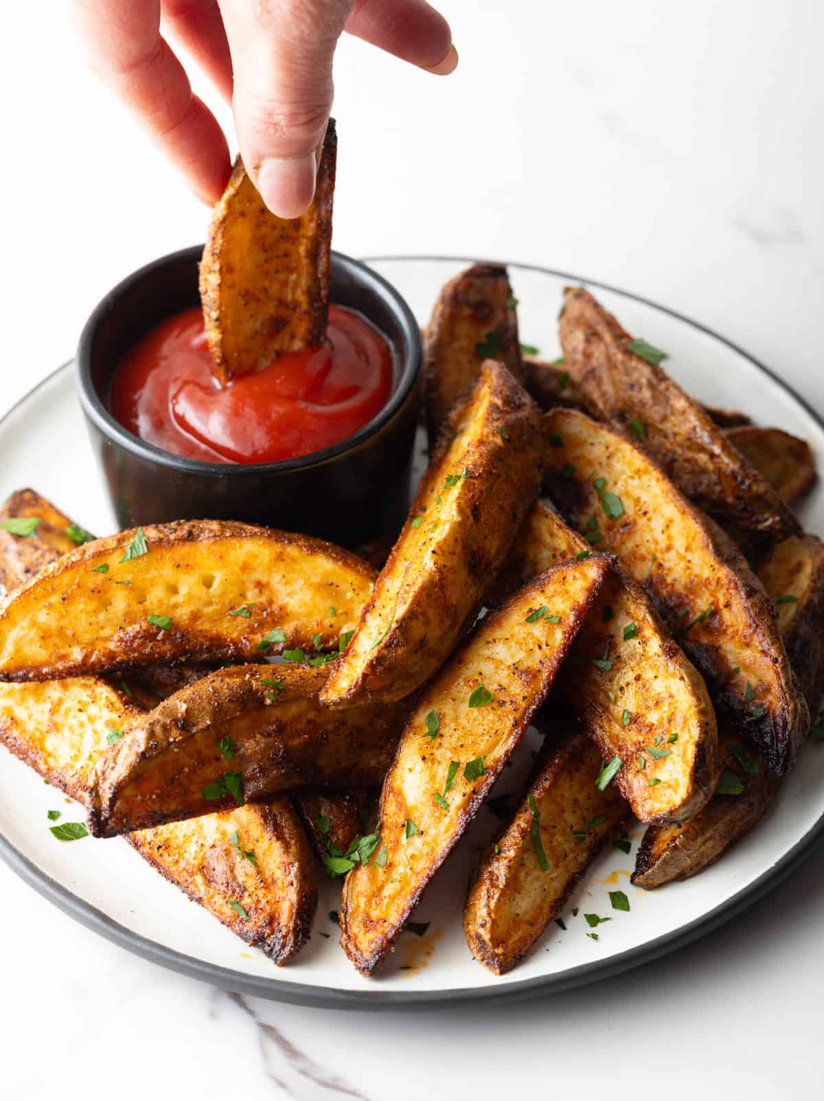 A plate full of oven baked potato wedges with a hand dipping one of the wedges into a ramekin of ketchup.