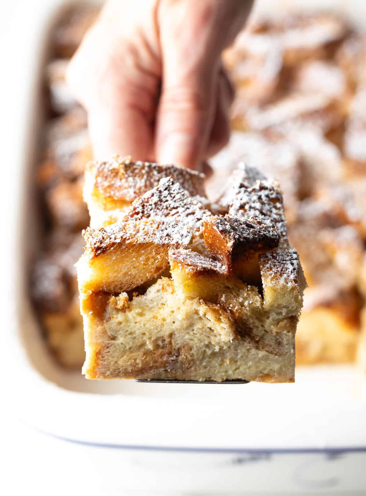 A slice of French toast casserole overnight recipe being held up with a spatula over a white baking dish.