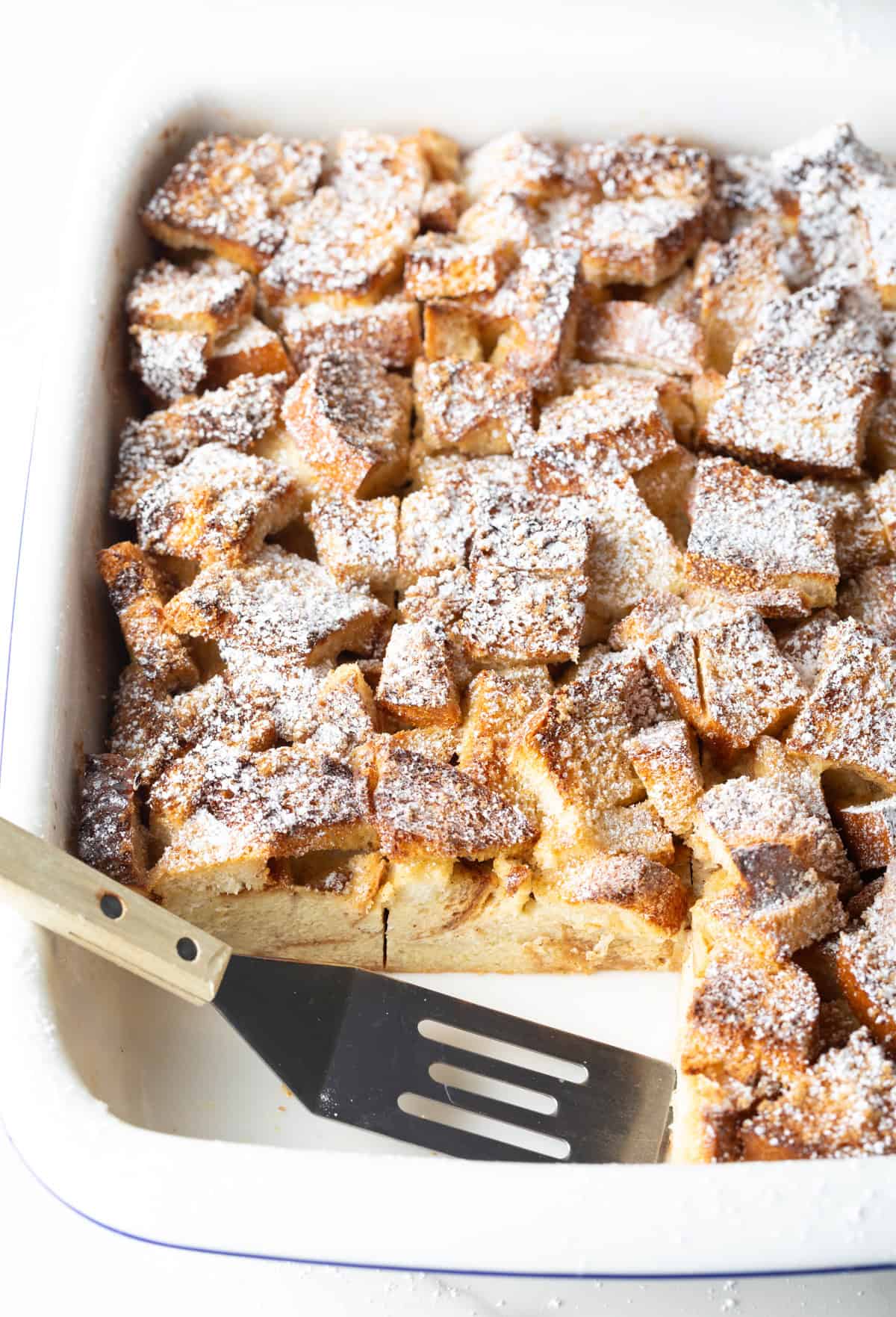 A white baking dish filled with overnight French toast breakfast casserole, with a metal spatula having taken two slices out.