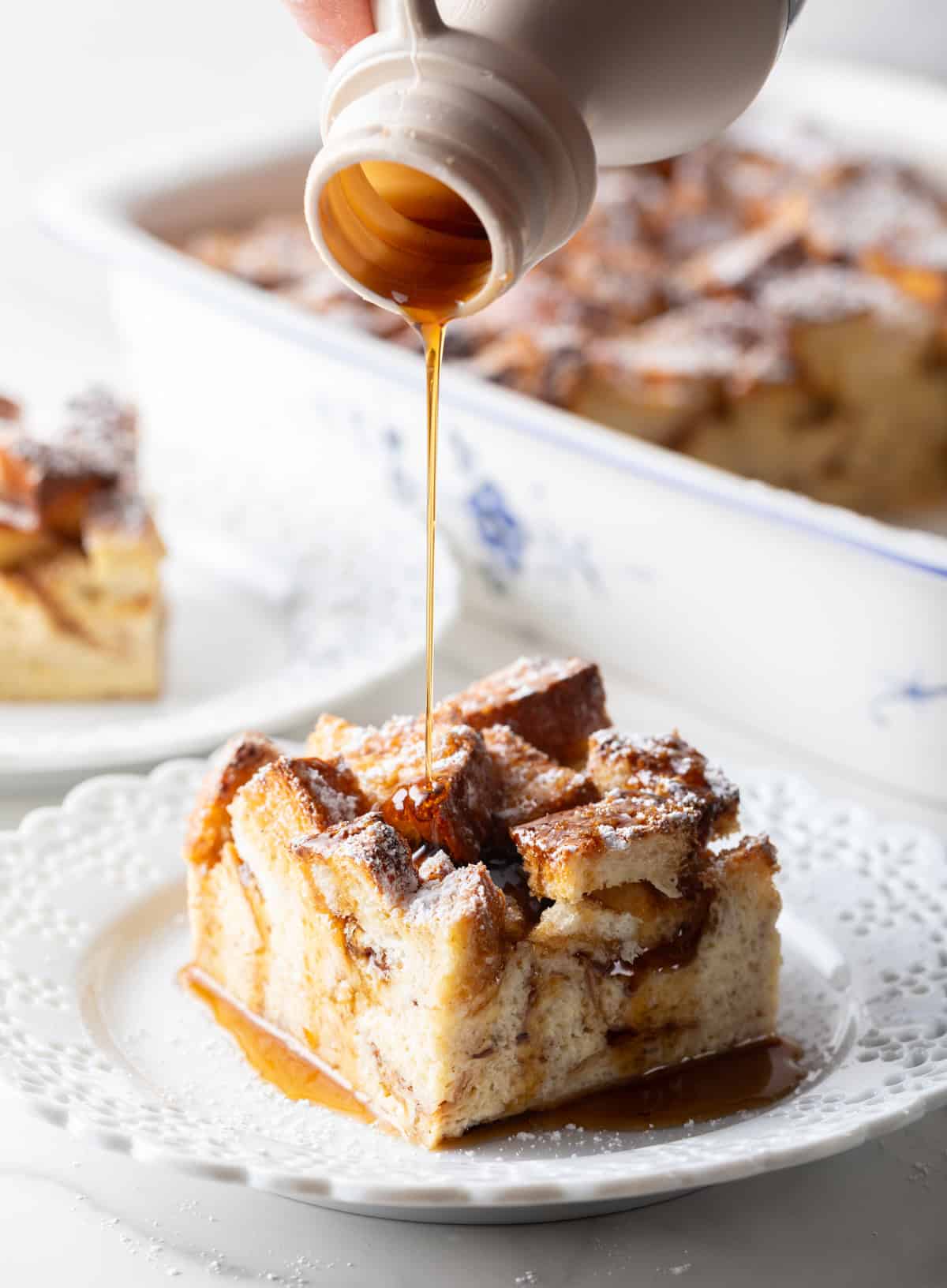 Syrup is being drizzled over a slice of make ahead French toast casserole on a white plate.