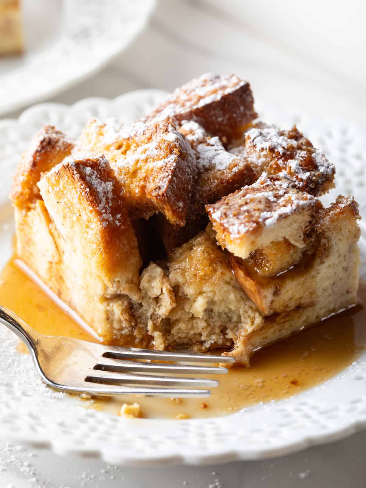 A serving of French toast casserole overnight recipe on a white plate drizzled with syrup, with a fork by its side.