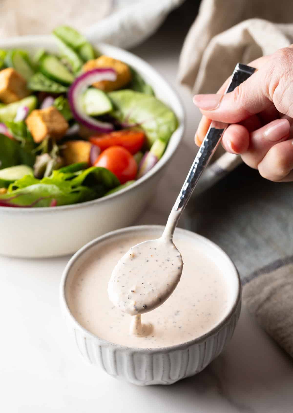A metal spoon is holding a serving of parmesan peppercorn dressing over a bowl of the dressing, next to a salad.