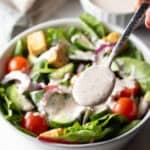 A metal spoon is ladling parmesan peppercorn dressing over a garden salad.