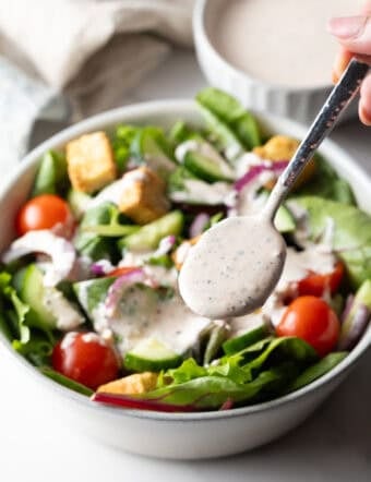A metal spoon is ladling parmesan peppercorn dressing over a garden salad.