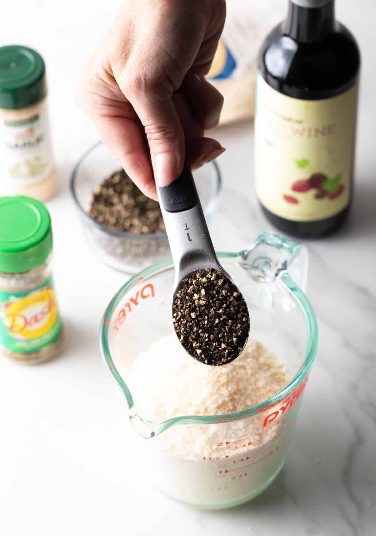 A measuring spoon is holding ground peppercorns over a mixture of parmesan cheese and mayonnaise.