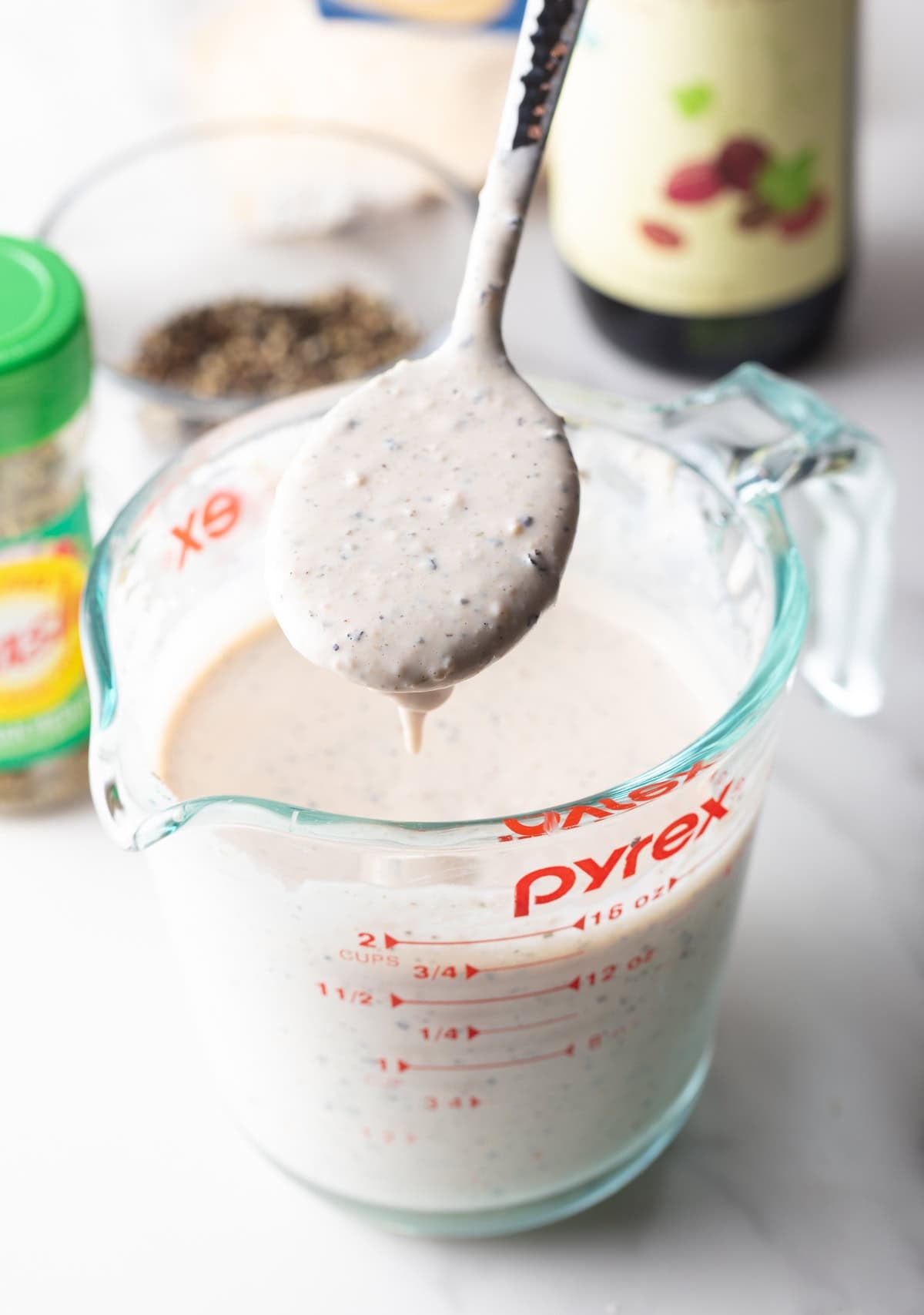 A spoon is holding fully mixed parmesan peppercorn dressing over a glass measuring cup.