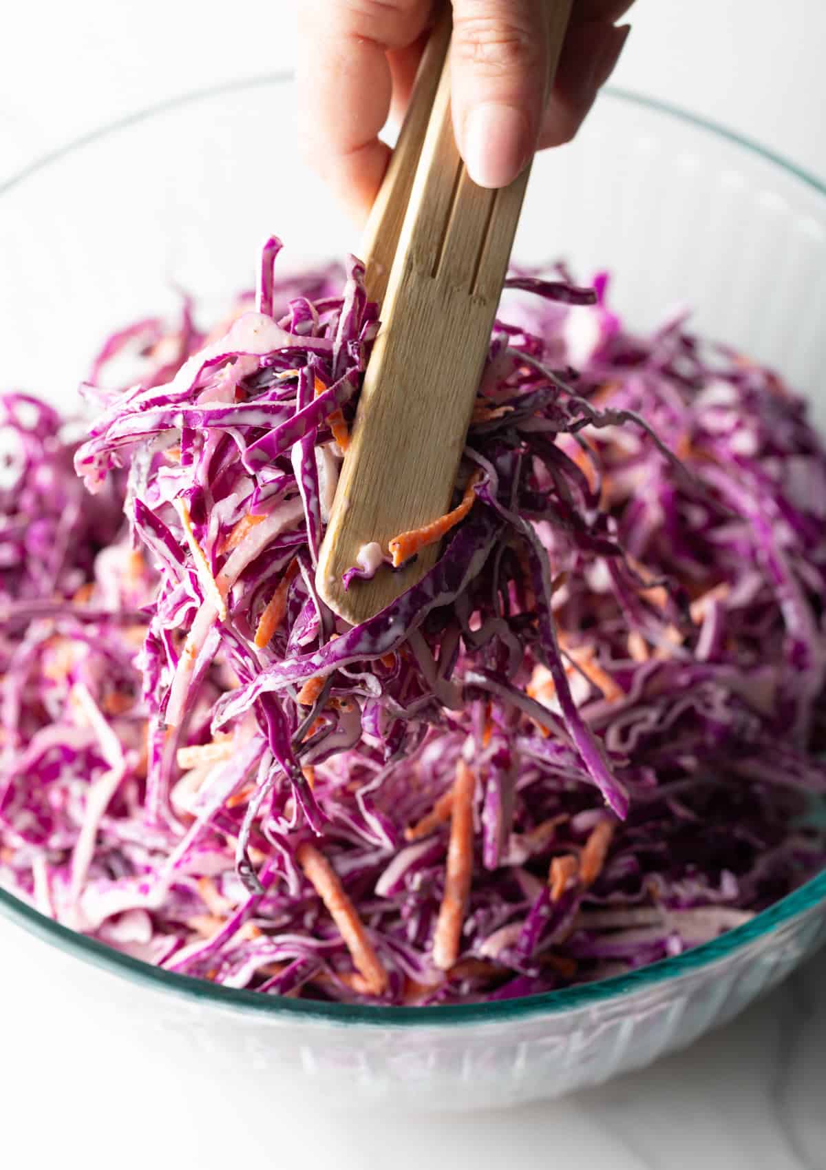 Red cabbage slaw is being tossed with wooden tongs in a glass bowl.
