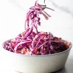 A fork is holding a bite of red cabbage slaw above a bowl of the cabbage slaw recipe.