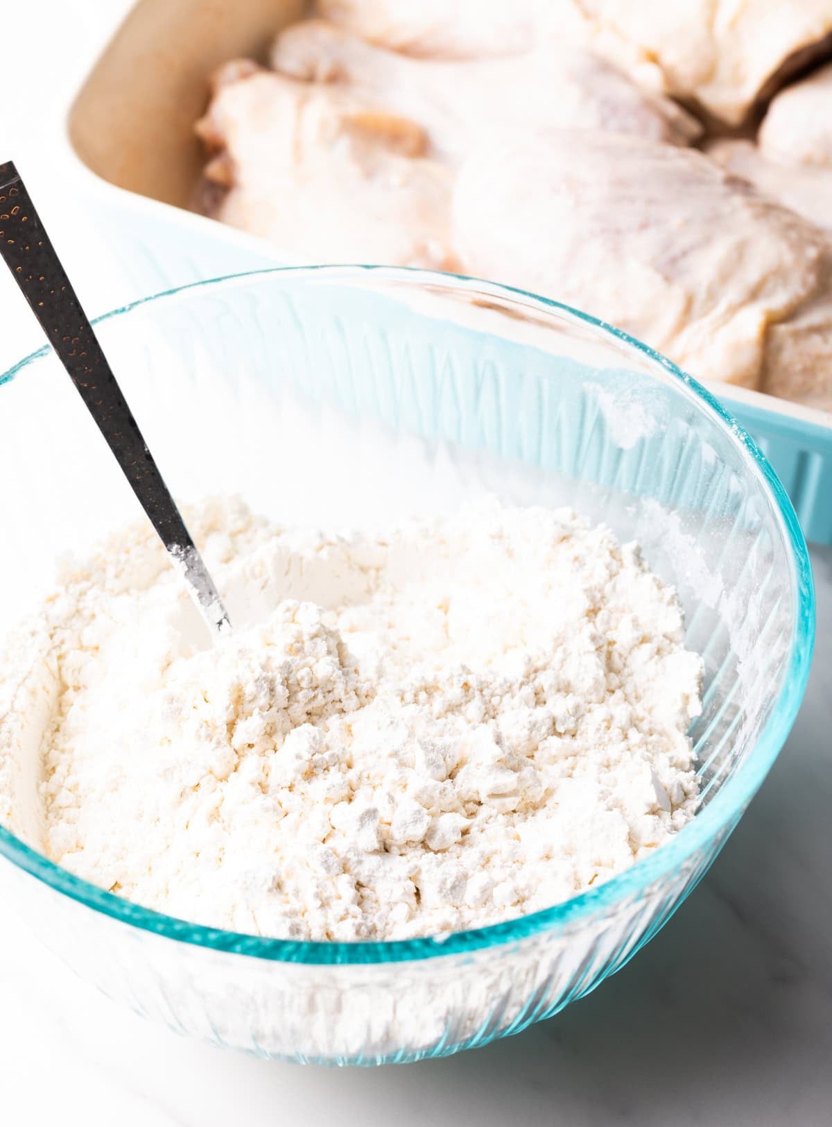 A spoon in a glass mixing bowl that is mixing a flour mixture for a deep fried chicken recipe.