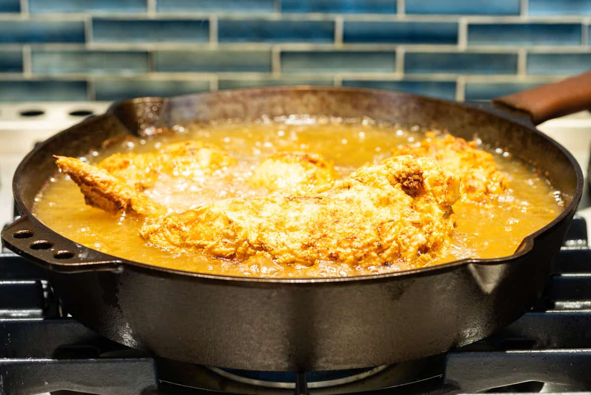 Fried chicken pieces are being cooked in a cast-iron skillet full of oil.