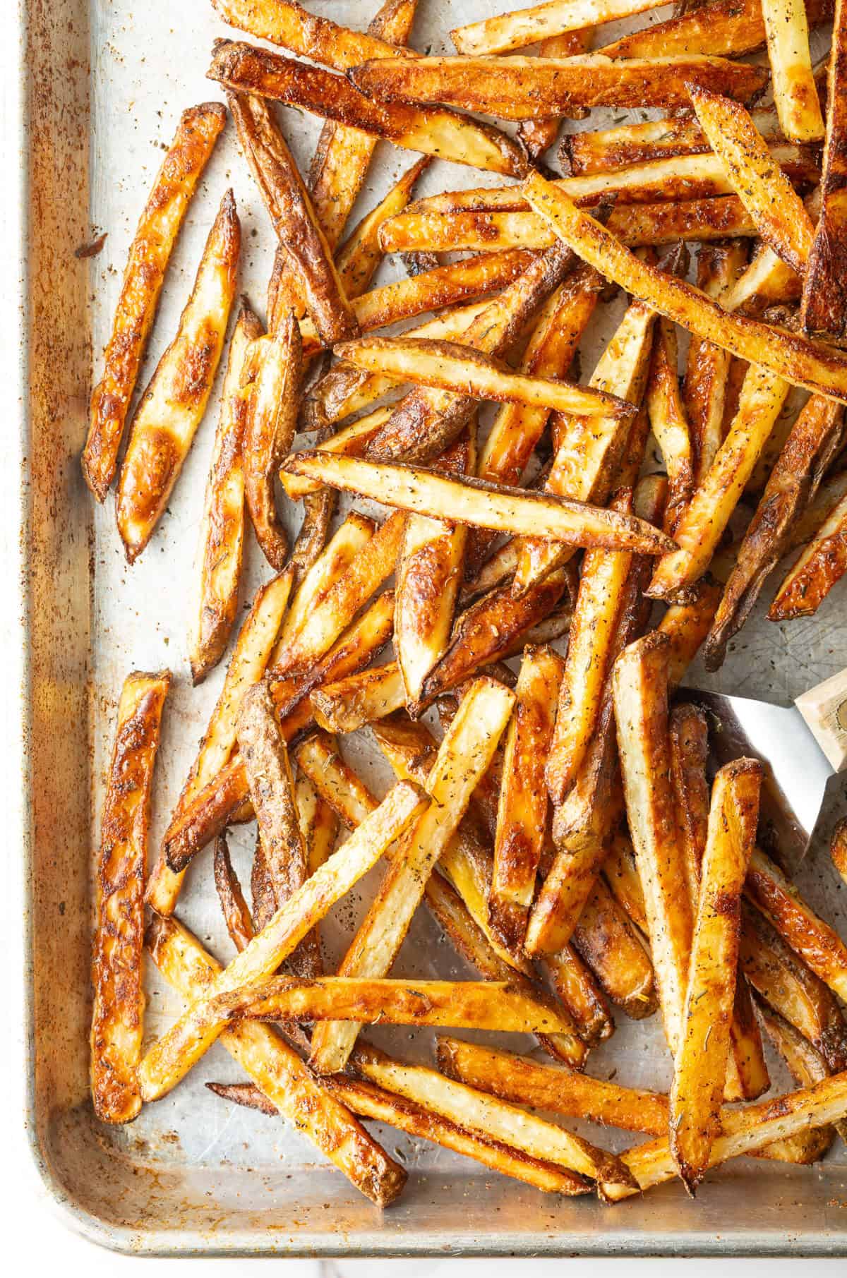 Crispy oven-baked French fries on a baking tray, being scooped by a metal spatula.