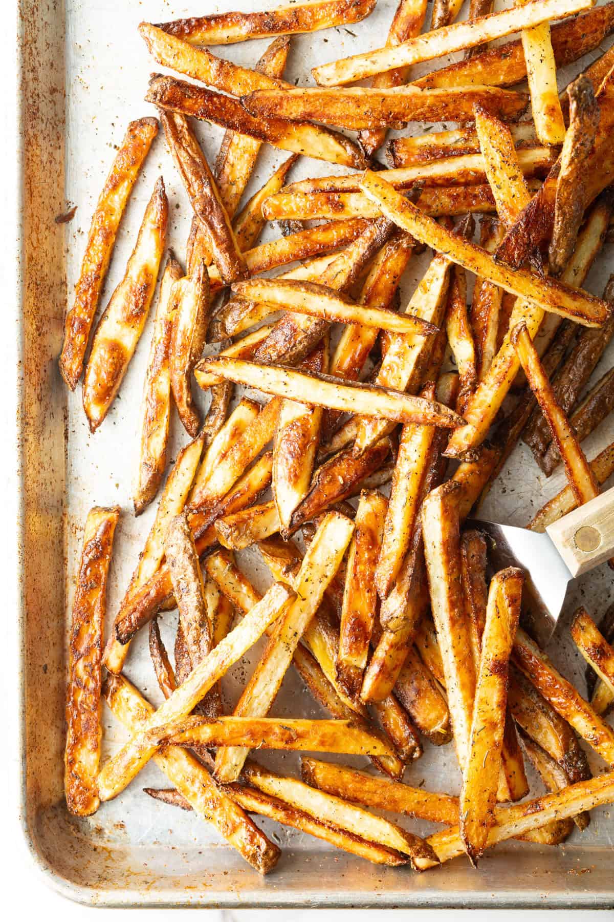 Oven baked French fries on a baking tray, being scooped by a metal spatula.