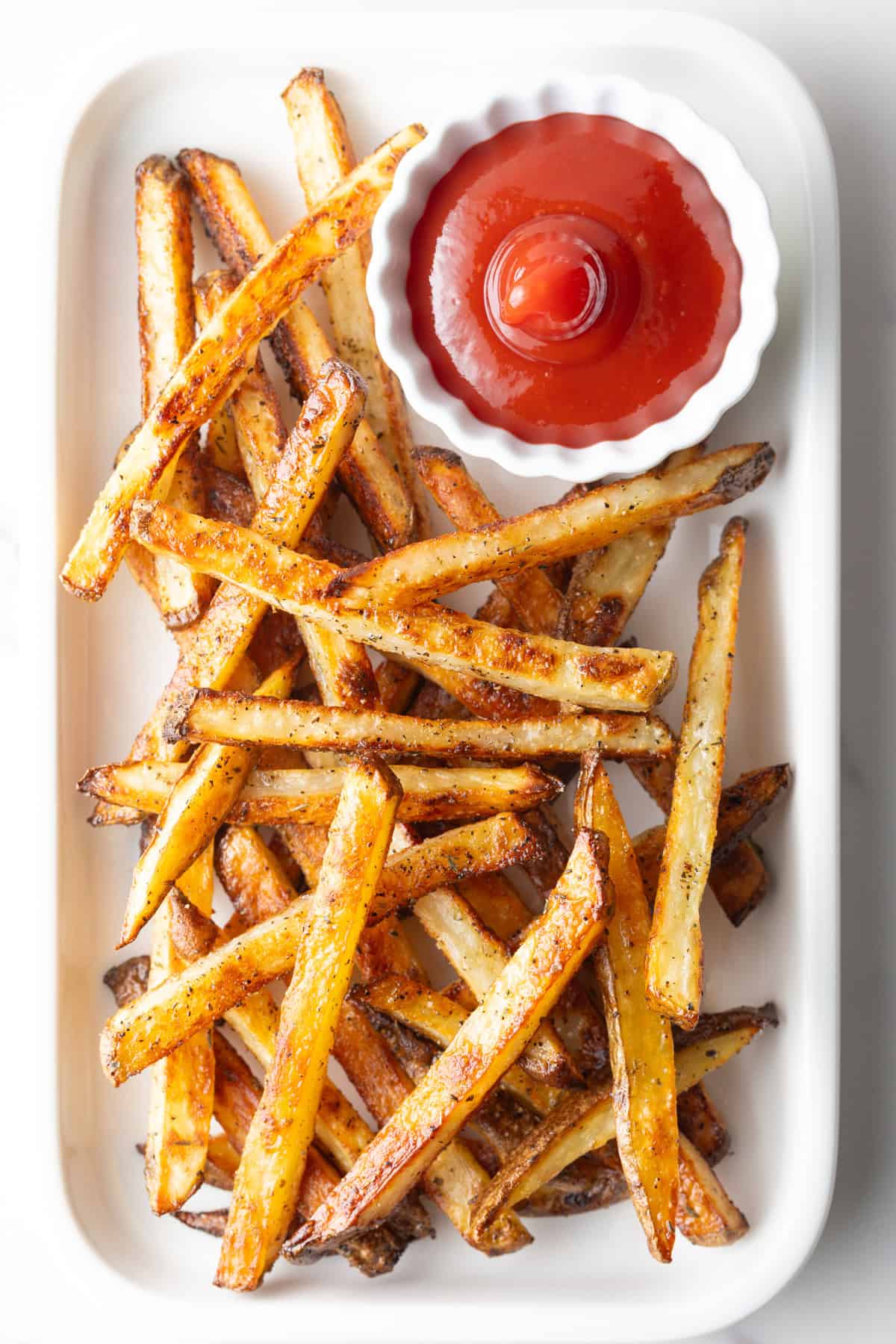 Homemade baked French fries from the oven sitting on a white plate with a ramekin of ketchup.