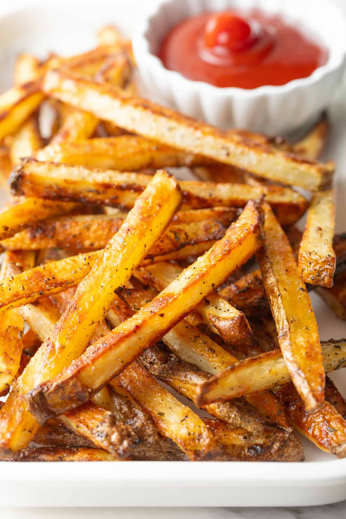 Homemade French fries made in the oven, sitting on a white plate next to a ramekin of ketchup.