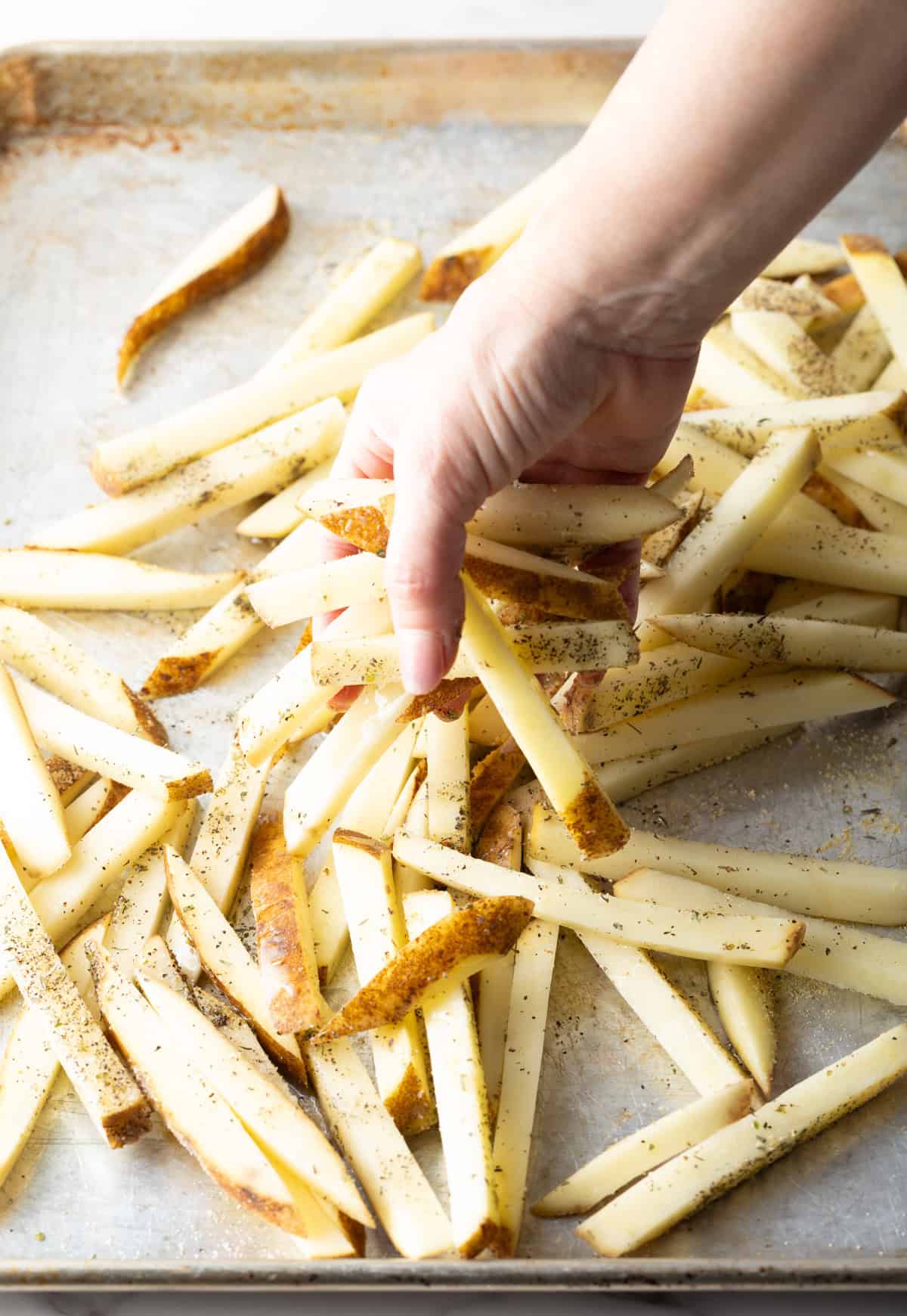 A hand is tossing raw potato slices in oil and seasoning, preparing them for a baked French fries recipe.