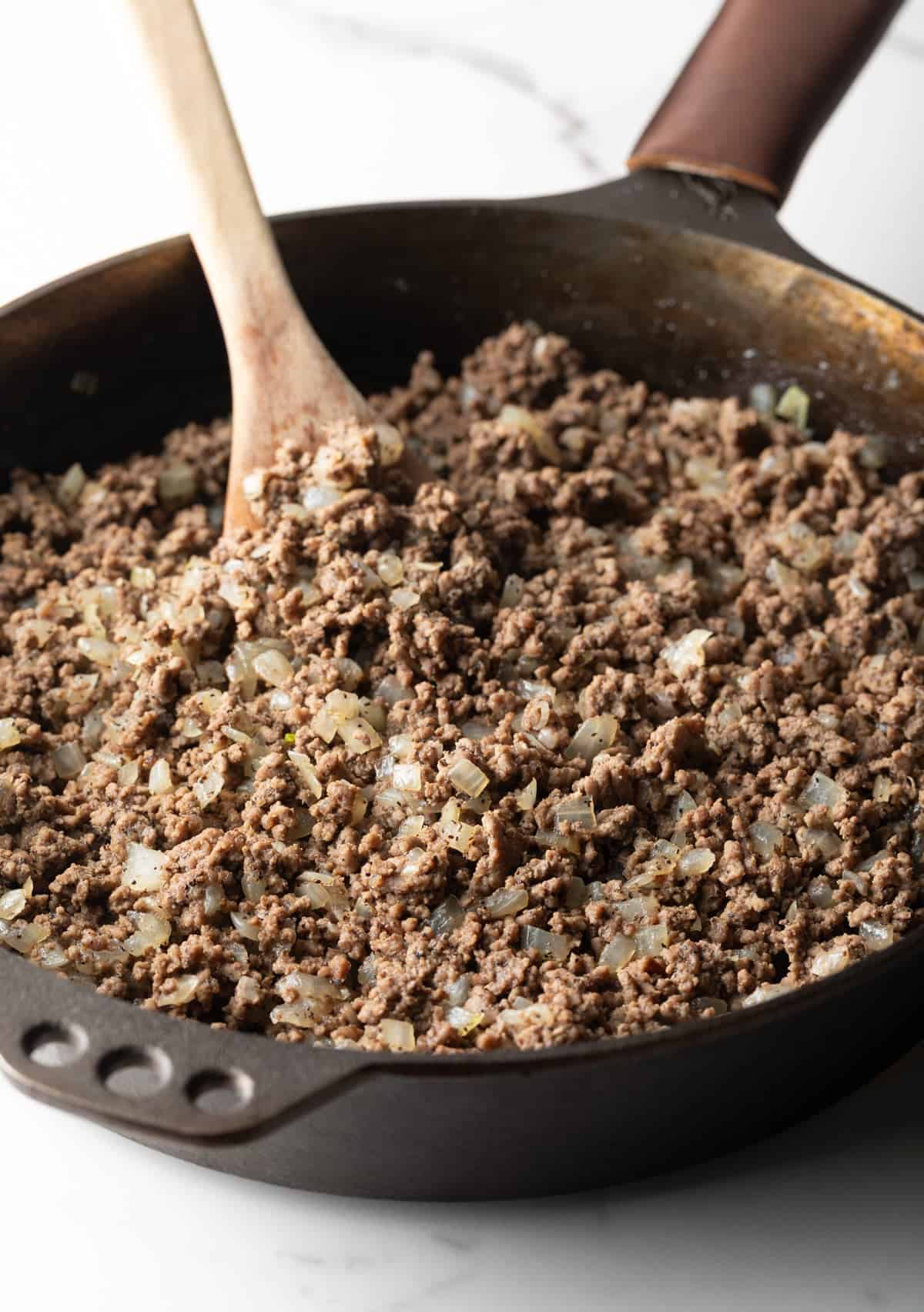 A beef and onion mixture is being cooked in a black skillet for an Amish hamburger casserole recipe.