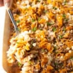 A metal spoon is holding a scoop of Amish hamburger noodle casserole above a dish filled with the casserole.