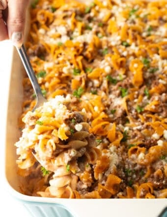 A metal spoon is holding a scoop of Amish hamburger noodle casserole above a dish filled with the casserole.