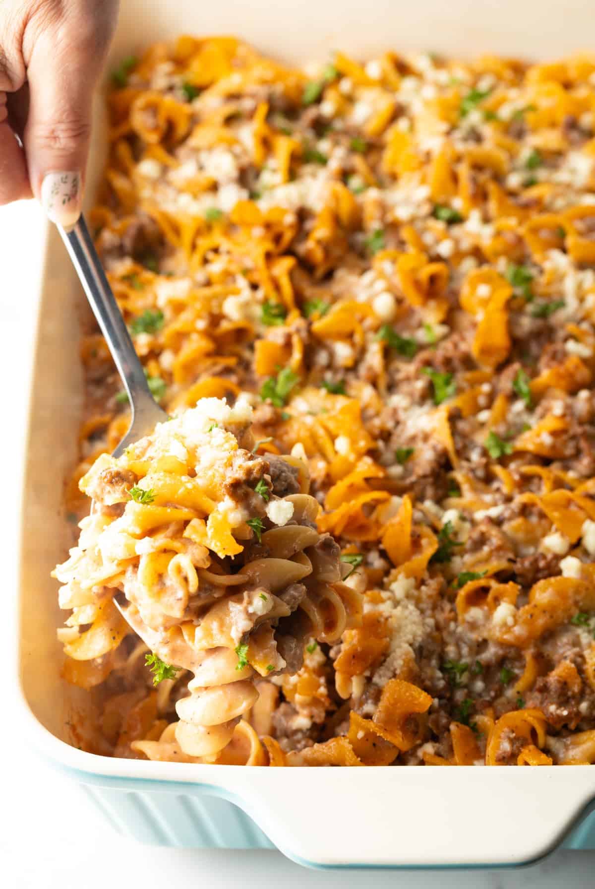 A metal spoon is holding a scoop of Amish hamburger noodle casserole above a dish filled with the casserole.