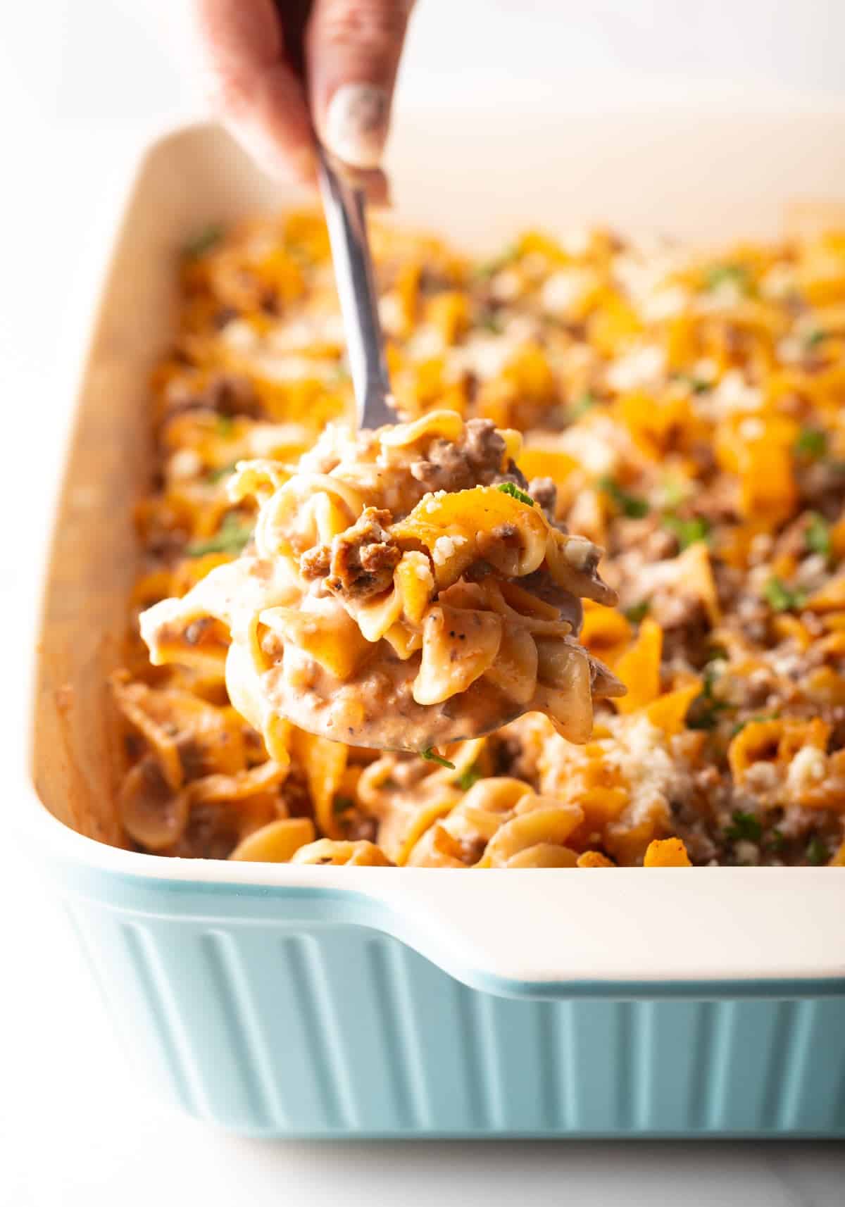 A metal spoon is holding a scoop of hamburger noodle casserole above a dish filled with the casserole.