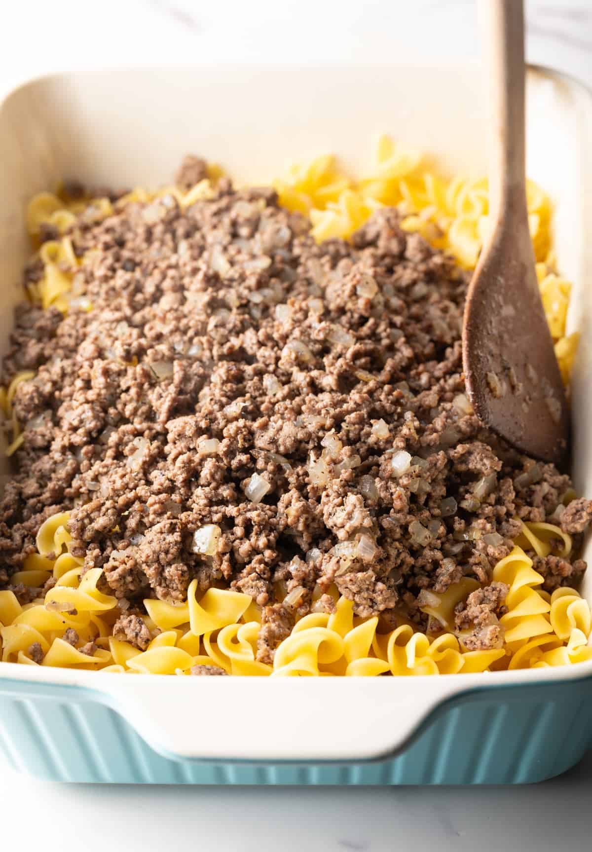 A ground beef mixture is being added over egg noodles in a white baking dish.