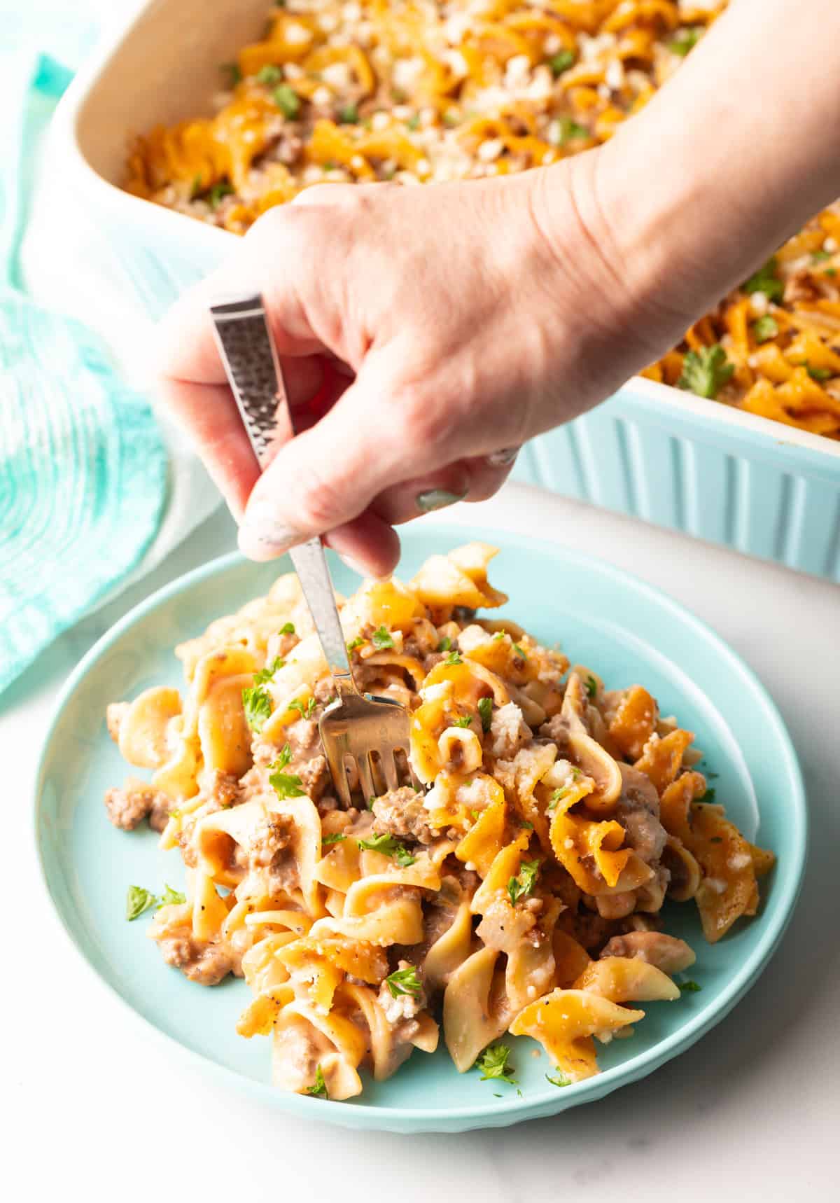 A serving of beef noodle casserole on a blue plate, with a hand holding a fork going for a bite.