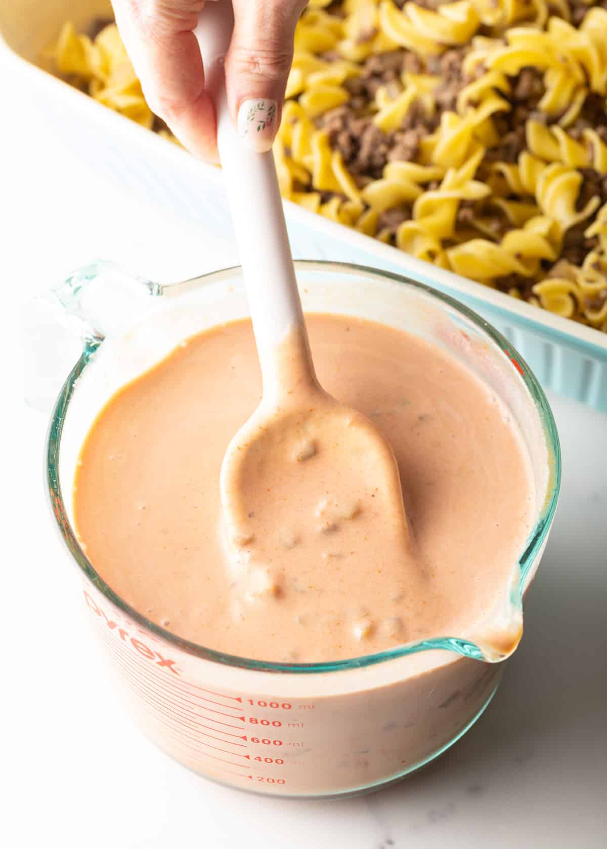 A creamy sauce mixture for a beef noodle casserole is being stirred in a glass measuring pitcher.