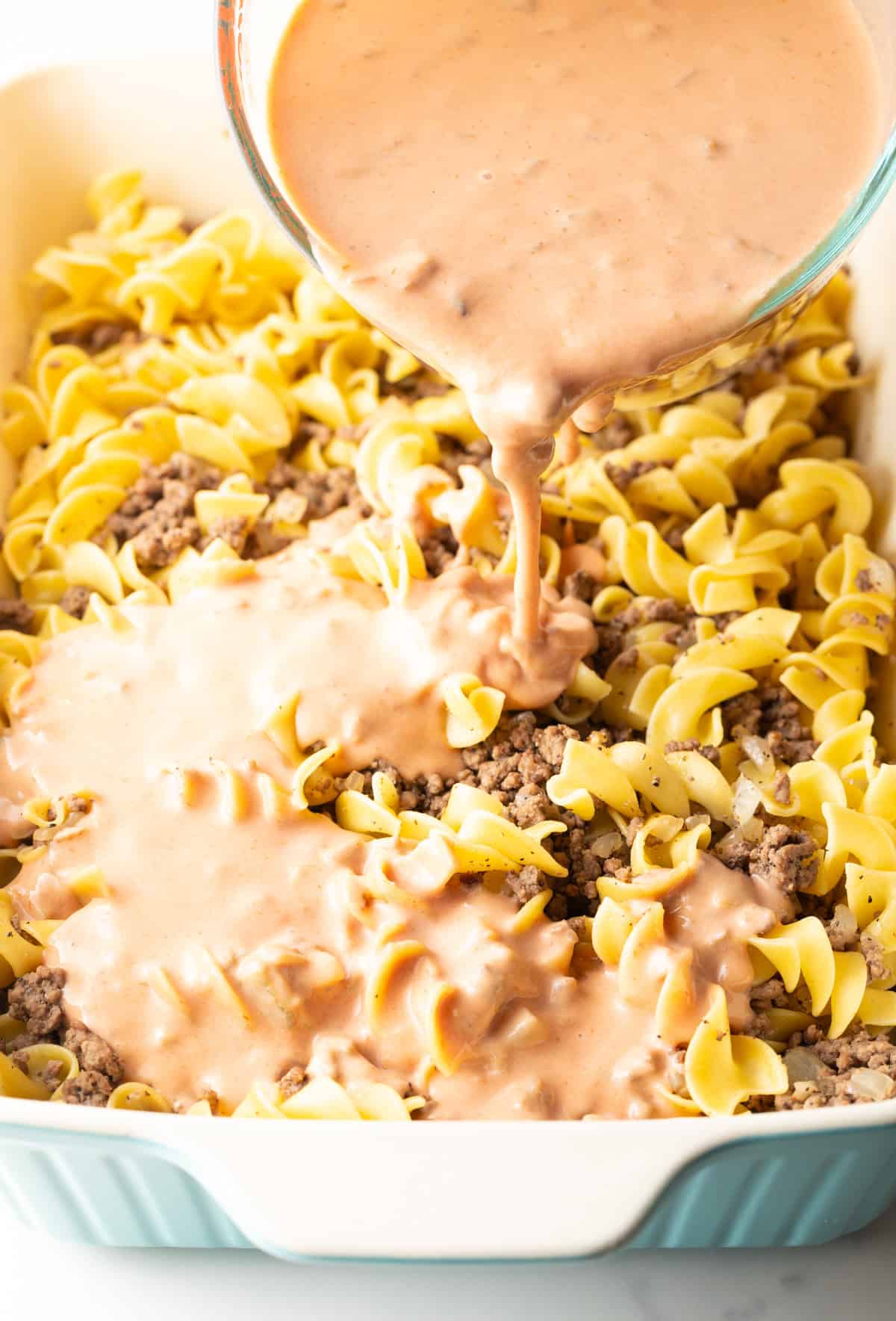 A cream mixture is being added to a baking dish filled with egg noodles and ground beef.