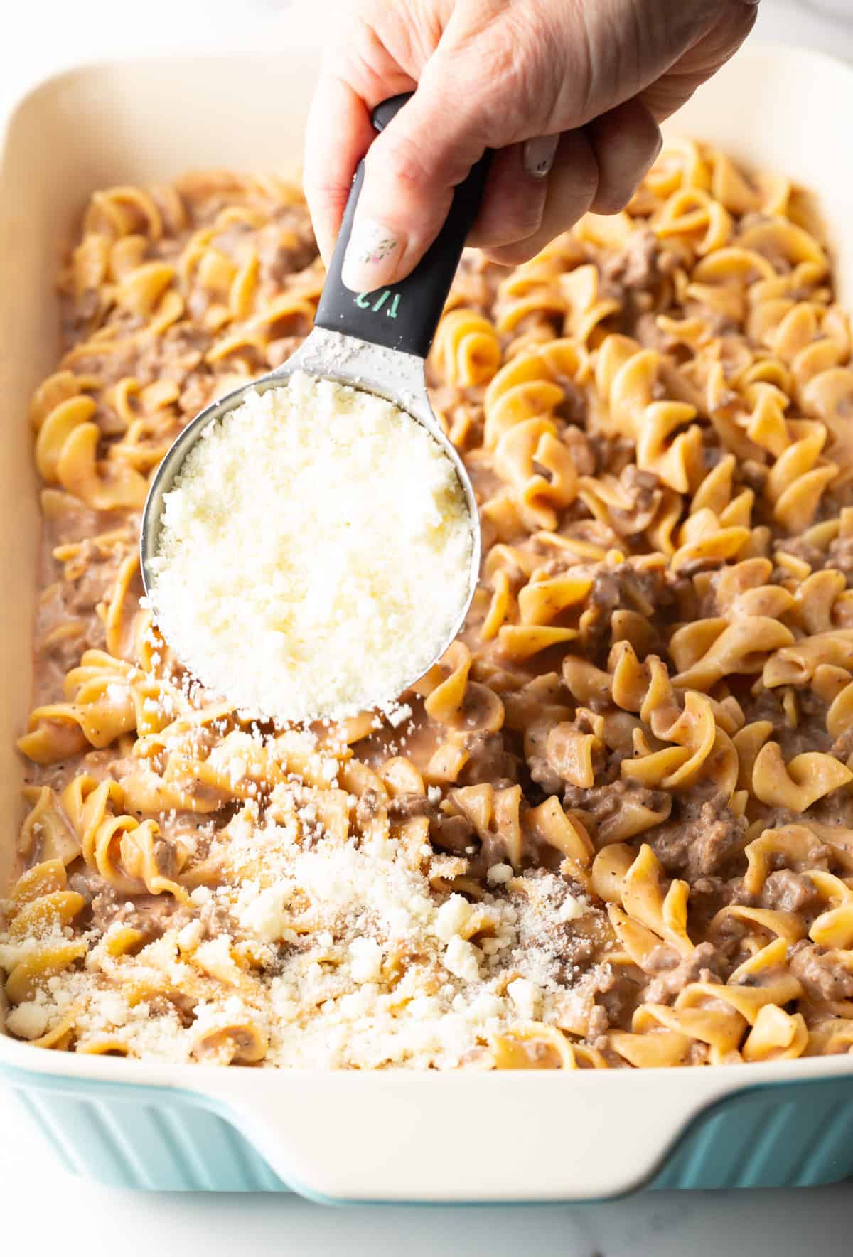 Shredded Parmesan cheese is being added on top of an Amish hamburger noodle casserole.