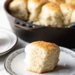 A fluffy homemade angel biscuit is sitting on a white plate, next to a skillet filled with angel biscuits.