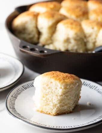A fluffy homemade angel biscuit is sitting on a white plate, next to a skillet filled with angel biscuits.