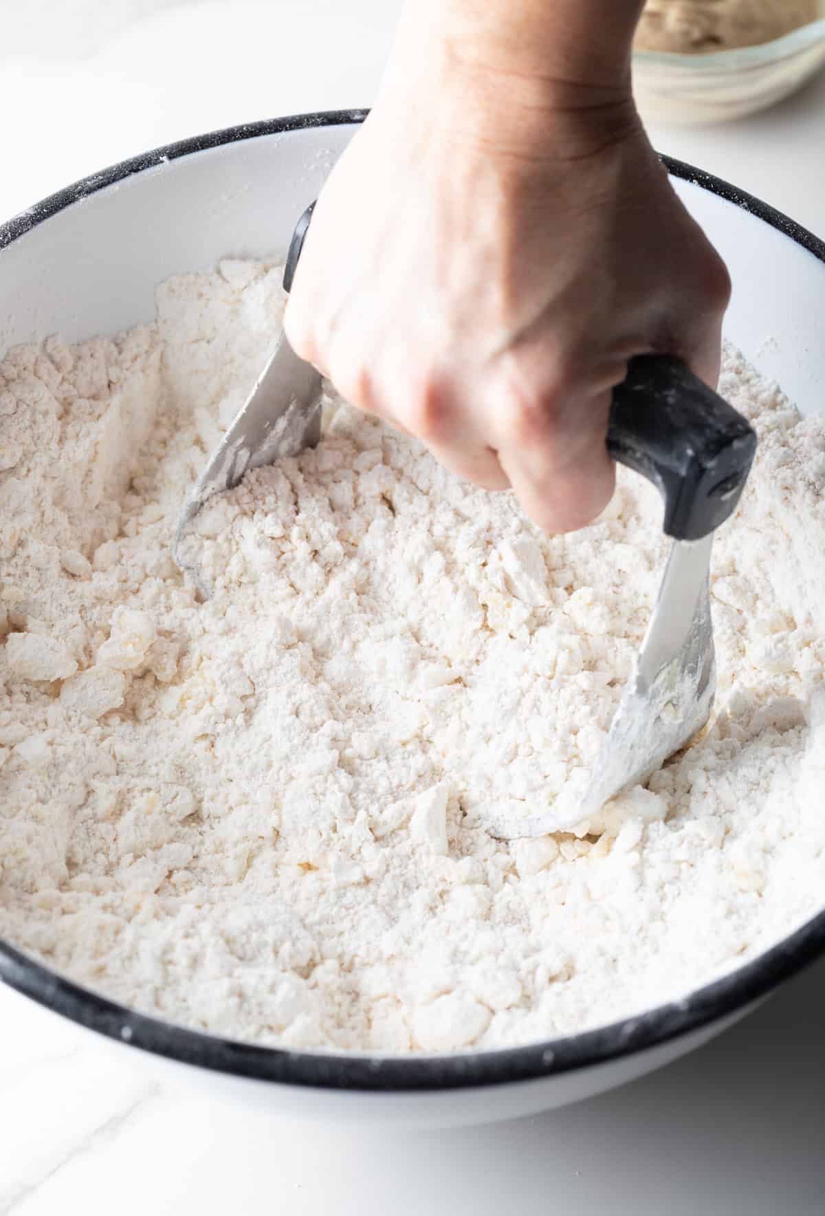 A hand holding a pastry cutter that is mixing butter and shortening into an angel biscuit mix.