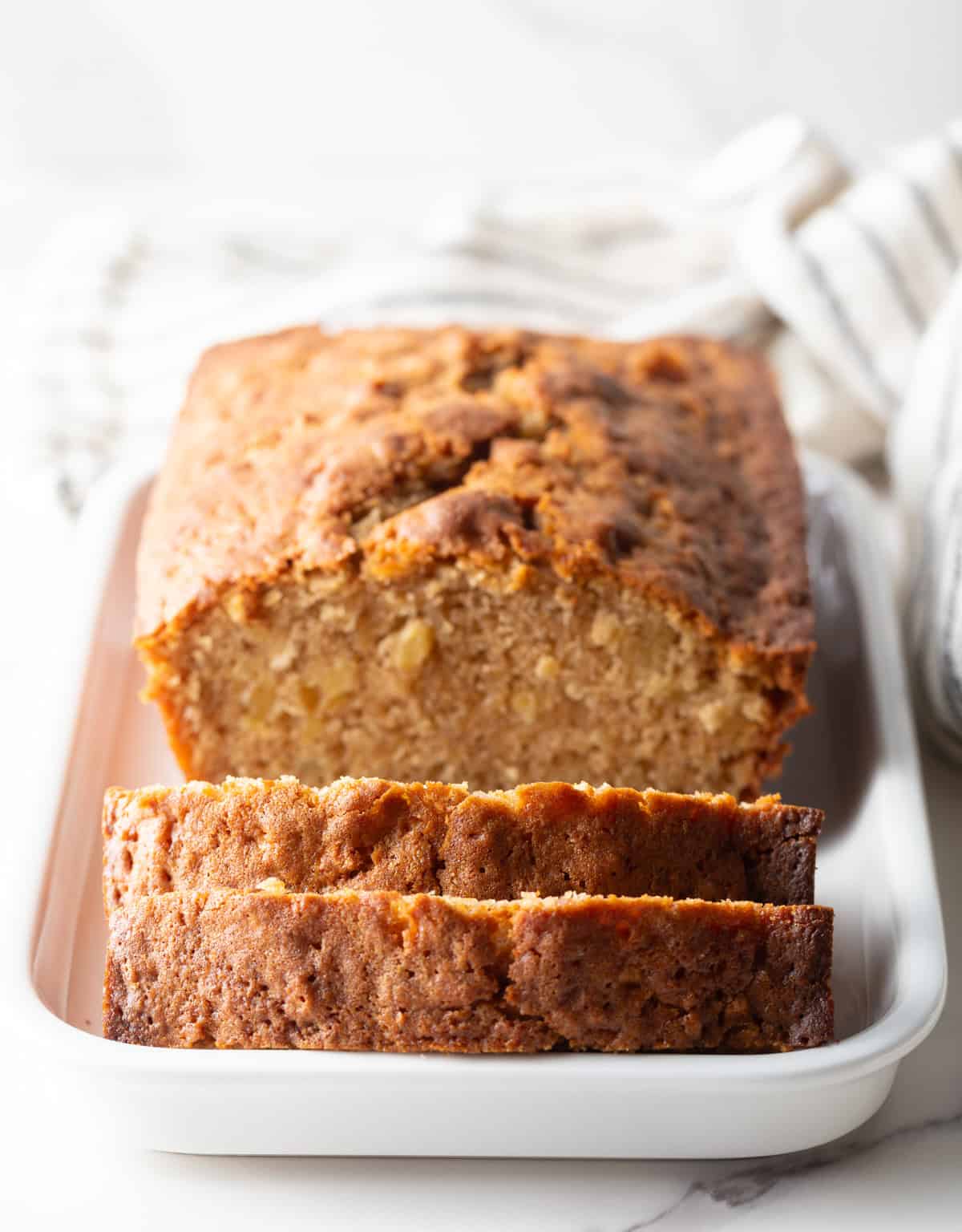 A freshly baked apple bread recipe sitting in a white baking dish, with a slice laying against the edge of the dish.