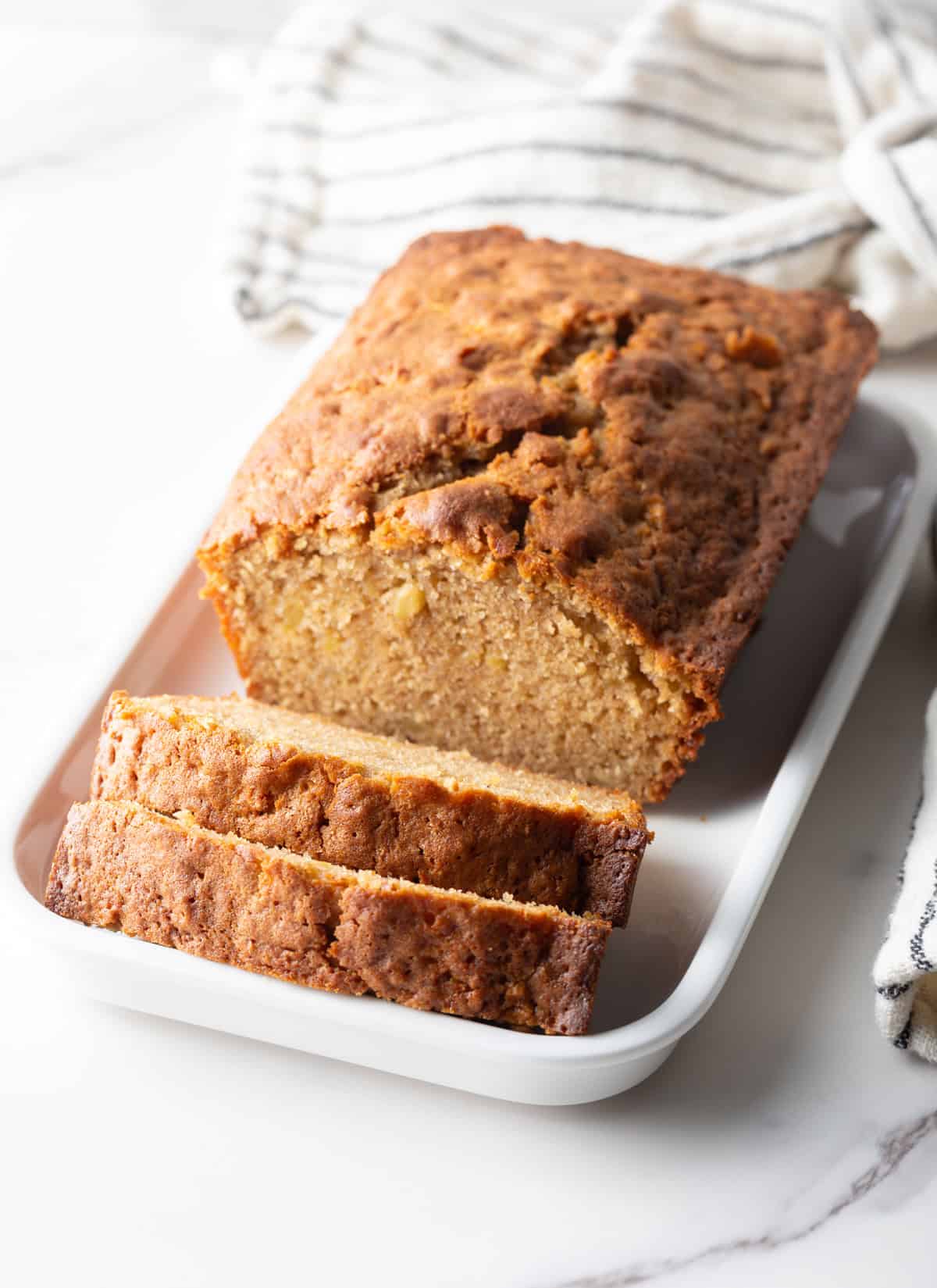 An apple bread recipe on a white dish with two slices cut in front of the loaf.
