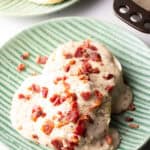 Two biscuits on a green plate, smothered with a bacon gravy recipe, with another plate of biscuits in the background.