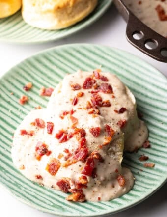 Two biscuits on a green plate, smothered with a bacon gravy recipe, with another plate of biscuits in the background.