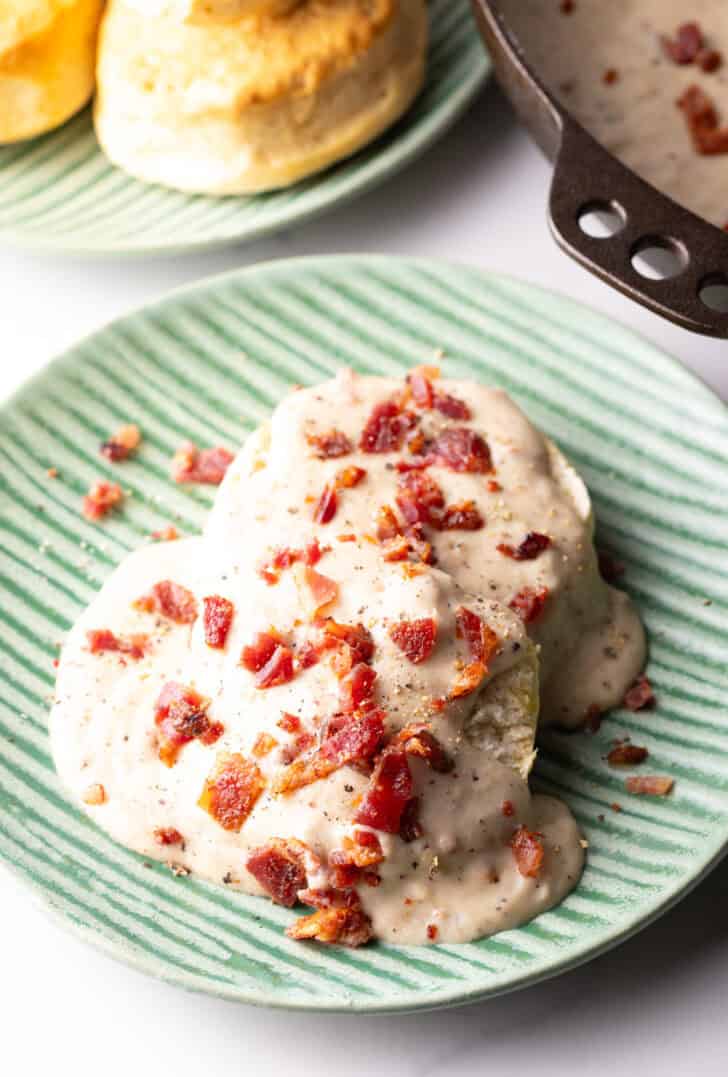 Two biscuits on a green plate, smothered with a bacon gravy recipe, with another plate of biscuits in the background.