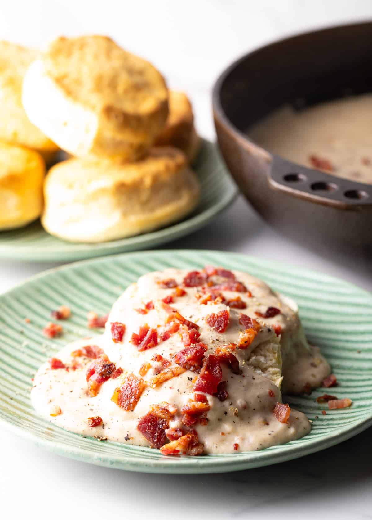 Two biscuits on a green plate, smothered with a bacon gravy recipe, with another plate of biscuits in the background.