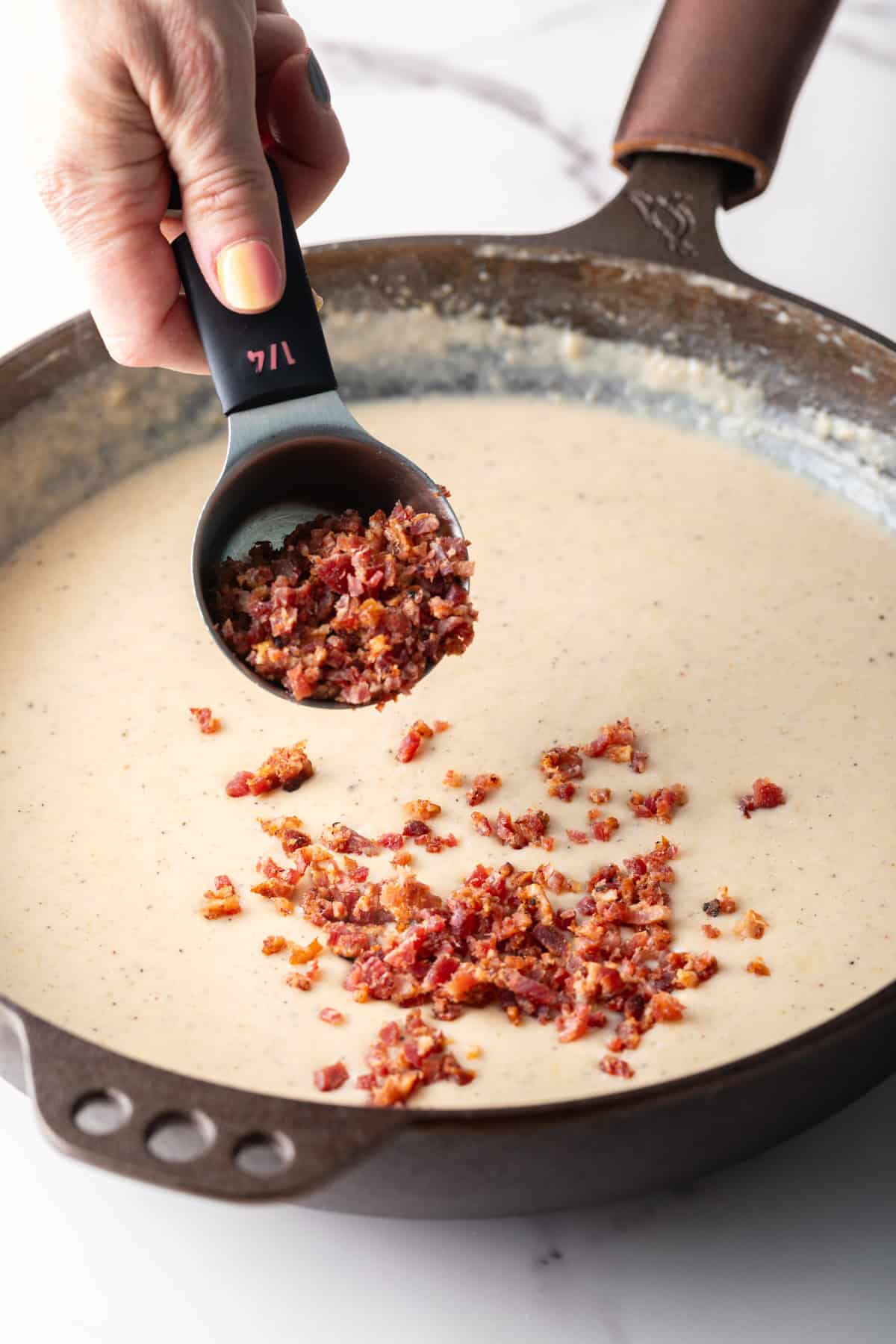 Crumbled bacon is being added to a bacon gravy recipe with a measuring cup.