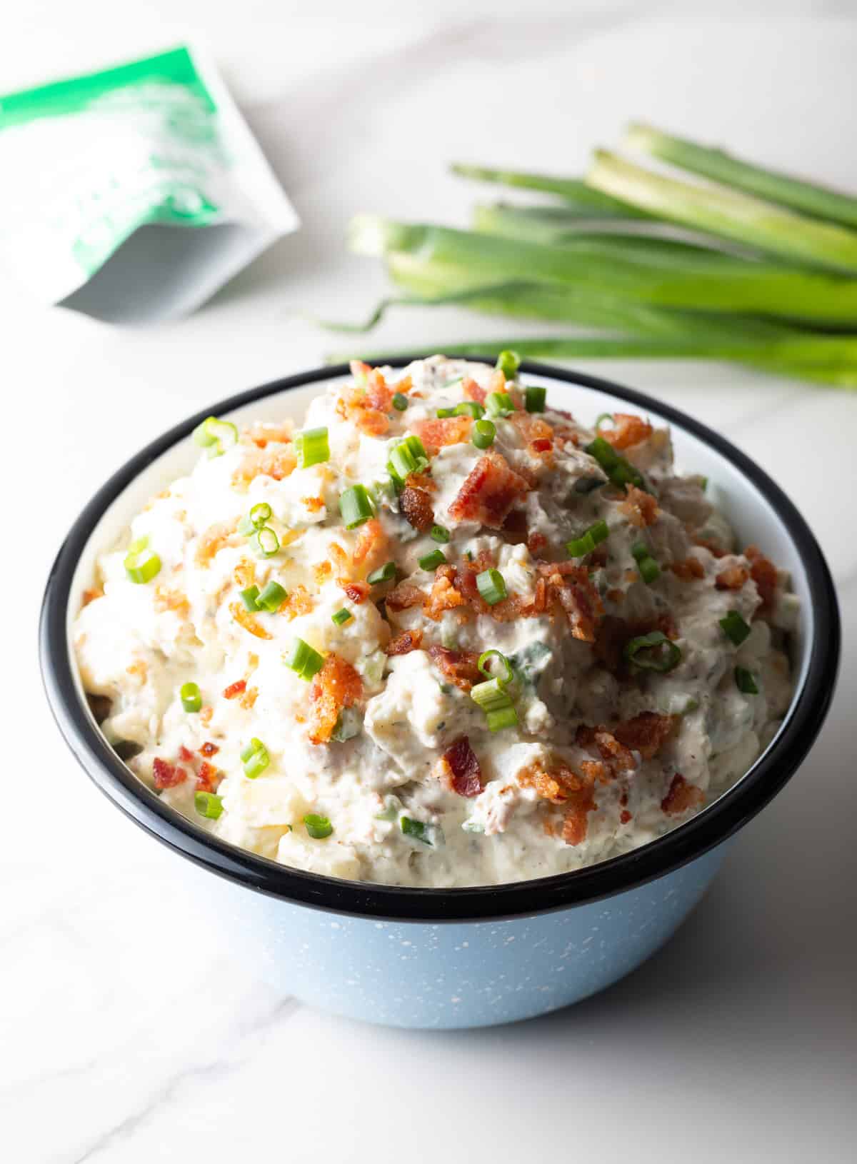 A blue bowl filled with a bacon ranch potato salad recipe in front of a bundle of green onions.